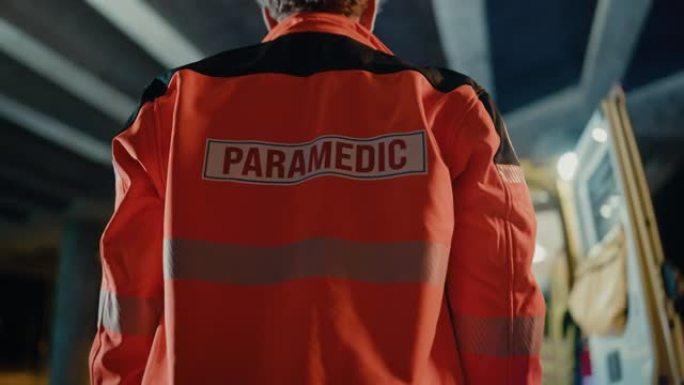 一名EMS护理人员的低弧镜头自豪地站在他的背上，穿着带有 “护理人员” 文字徽标的高可见度医用橙色制