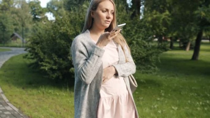 年轻怀孕的金发女郎走在城市公园用智能手机录制音频信息