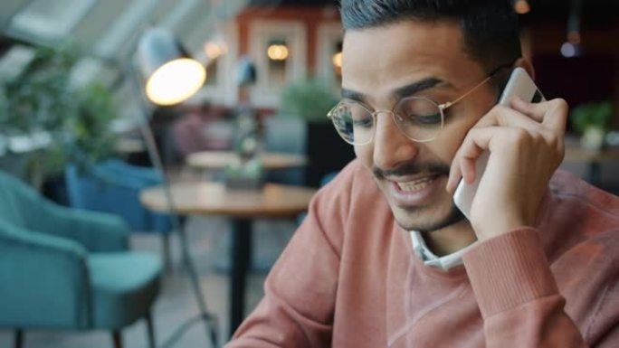 快乐的阿拉伯男人在手机上聊天并在现代咖啡馆做笔记的肖像