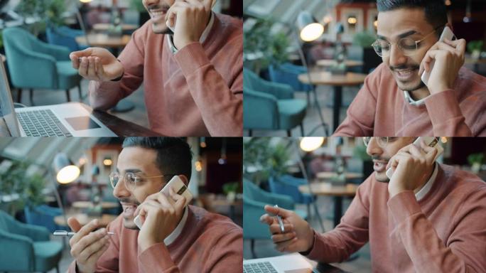 快乐的阿拉伯男人在手机上聊天并在现代咖啡馆做笔记的肖像