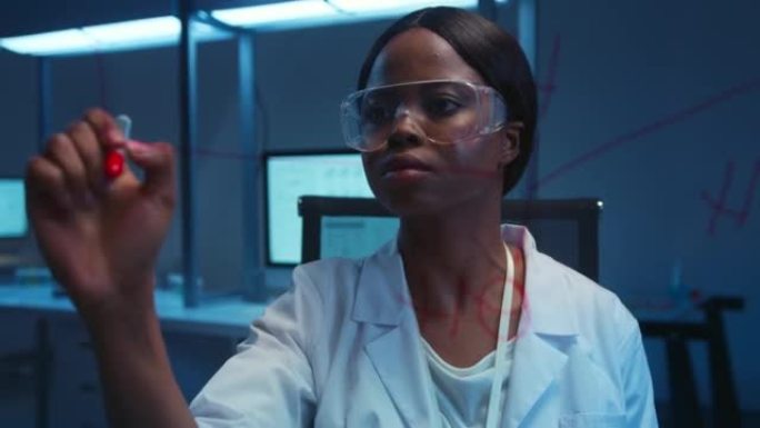 一位年轻的深色皮肤的实验室助手戴着防护眼镜，并在实验室的玻璃板上写了一件实验服