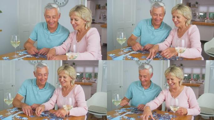 高级退休夫妇坐在家里的餐桌旁，端着一杯葡萄酒做拼图游戏
