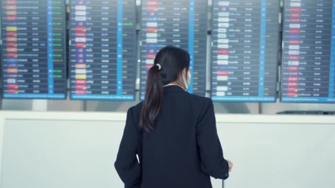 亚洲女商人戴着口罩，手提箱站在木板前，看信息检查她在国际机场的航班。