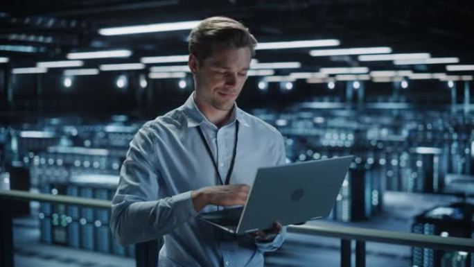 在数据中心使用笔记本电脑的英俊微笑的IT专家。成功的商人和电子商务企业家俯瞰服务器场云计算设施。中宽