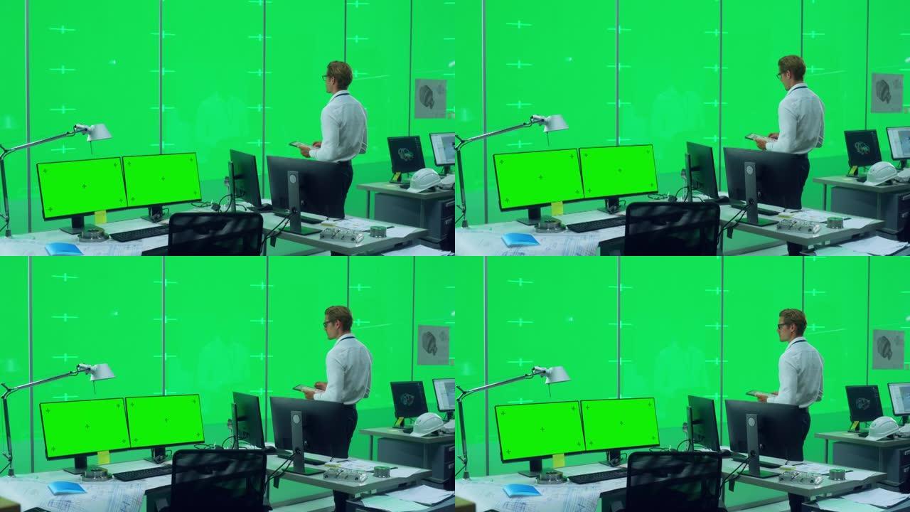 英俊的男工程师站在一个办公空间里，在一个绿色的虚拟屏幕色度键墙前有台式电脑，墙上有用于模型和模板的占