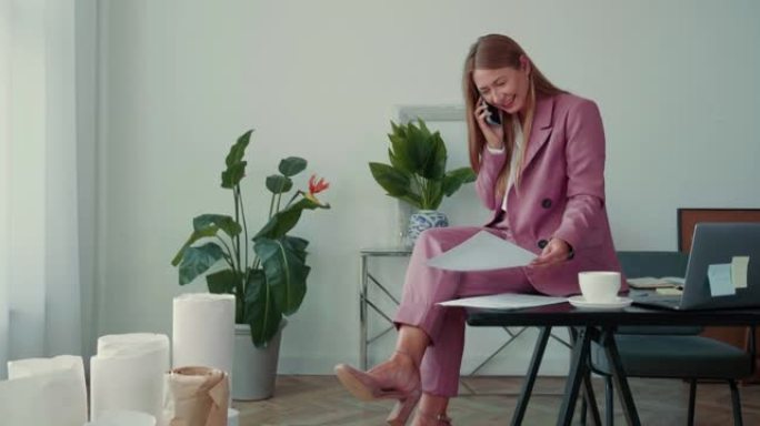 快乐成功的漂亮老板穿着粉色西装的女商人坐在办公室工作场所的桌子上通过手机交谈。