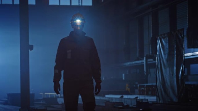 专业的重工业工程师/工人穿着制服，手电筒在钢铁厂的安全帽上。工业专家在深色金属建筑制造商中走向相机。