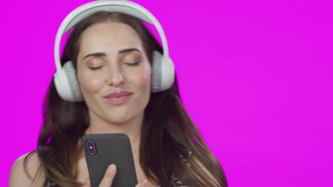 4k视频片段，一个美丽的年轻女子使用带智能手机的耳机，在粉红色的工作室背景下跳舞