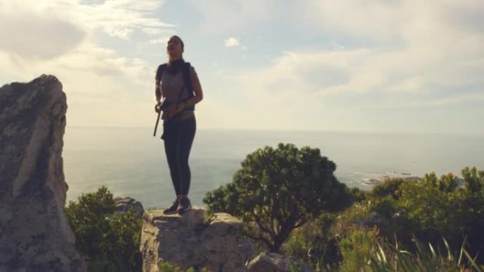 适合活跃的女人使用双筒望远镜在山上徒步旅行。徒步旅行者穿着背包站在悬崖边，在大风天看着背景为日落和海