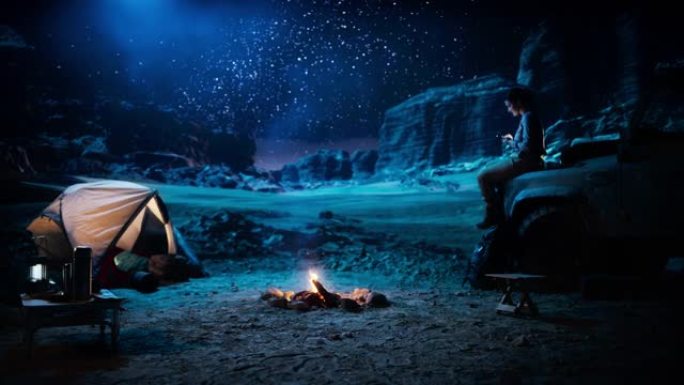 坐着的女性旅行者使用智能手机，在篝火旁在峡谷中露营时凝视着星空。自信的女冒险家在社交媒体互联网上分享
