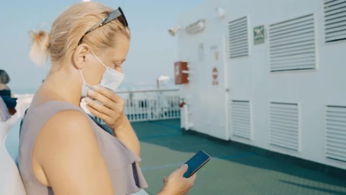带着智能手机的年轻女子在船甲板上戴上口罩