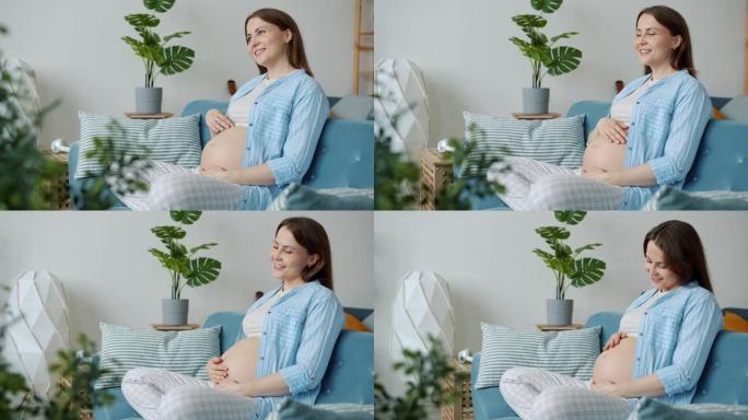 迷人的孕妇母亲坐在家里的沙发上抚摸肚子的肖像