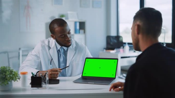 非裔美国医生正在健康诊所的绿屏计算机上向患者解释诊断。穿着白大褂的助理正在医院办公室阅读病史。