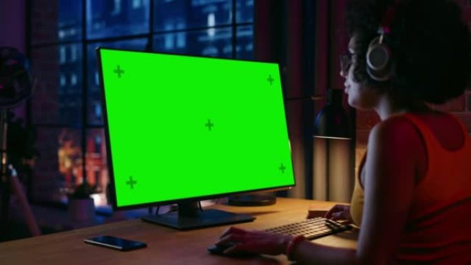 戴着耳机的年轻漂亮女性在台式电脑上在家工作，绿屏模拟显示。有创造力的女人检查社交媒体，浏览互联网。从
