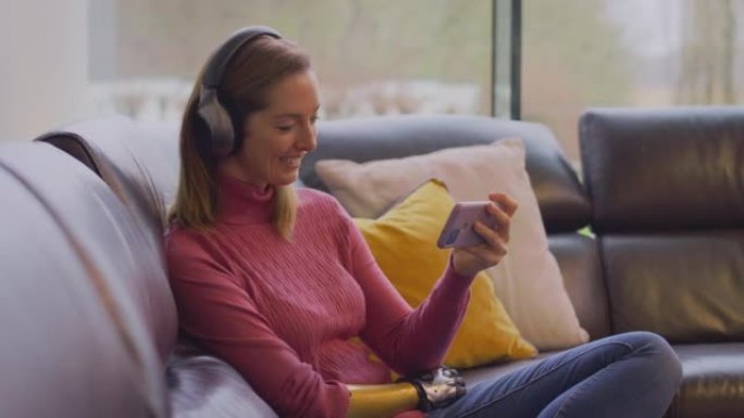 女人戴着无线耳机，在家中坐在沙发上听手机上的音乐流-慢动作拍摄