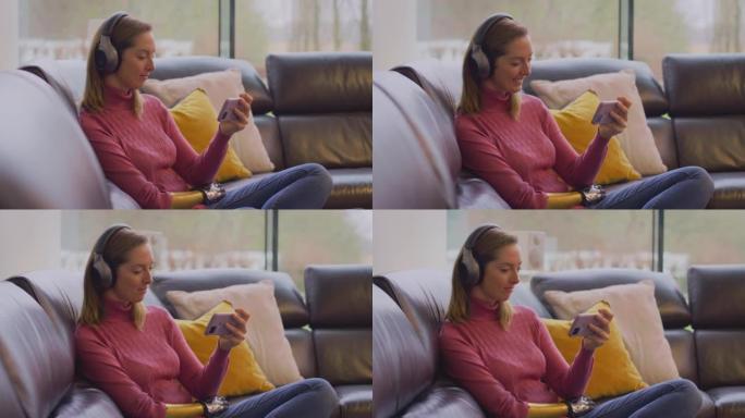 女人戴着无线耳机，在家中坐在沙发上听手机上的音乐流-慢动作拍摄