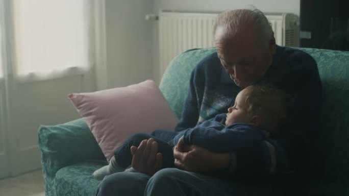电影拍摄的温柔的高级白发祖父正在亲吻他的孙子婴儿，坐在家里的沙发上安静地睡在手臂上。概念: 生活，祖
