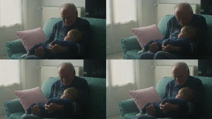 电影拍摄的温柔的高级白发祖父正在亲吻他的孙子婴儿，坐在家里的沙发上安静地睡在手臂上。概念: 生活，祖