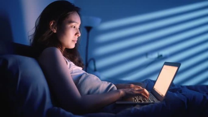 年轻的亚洲妇女工作到很晚，在床上使用笔记本电脑