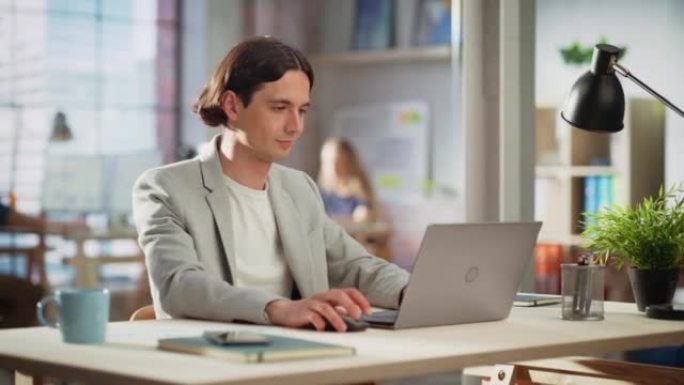 英俊的经理坐在创意办公室的办公桌前。穿着夹克的年轻时尚男人在营销机构使用笔记本电脑。在后台工作的同事