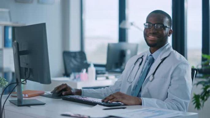 戴眼镜的非裔美国家庭医生正在一家健康诊所的电脑上工作。穿着白色实验室外套的成功黑人医生看着相机，在医