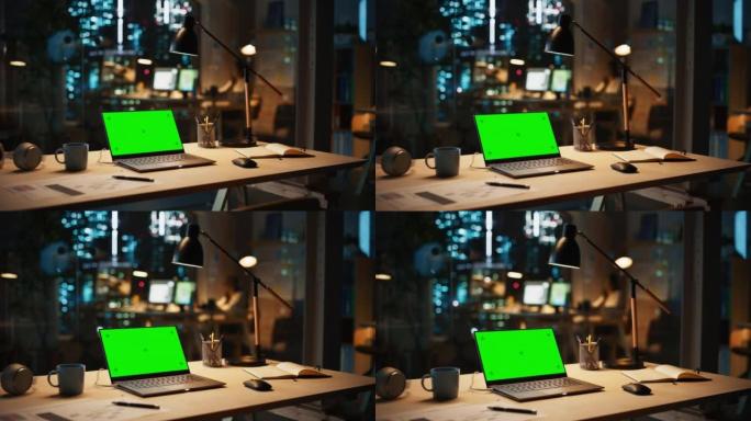 笔记本电脑站在桌子上，晚上在现代创意办公室里有一个绿屏Chromakey模拟显示器。员工在背景温暖的