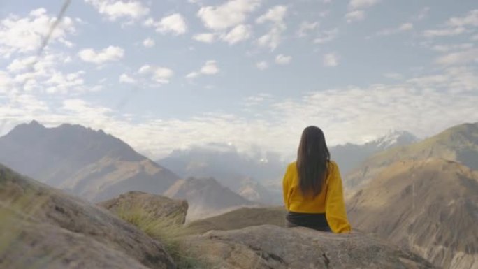 自由快乐的旅行者穿着黄色冬季夹克的年轻女性坐在岩石上，早上在山顶上享受旅行假期。种族多样化的旅行者概