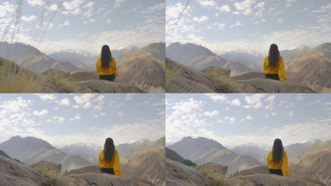 自由快乐的旅行者穿着黄色冬季夹克的年轻女性坐在岩石上，早上在山顶上享受旅行假期。种族多样化的旅行者概