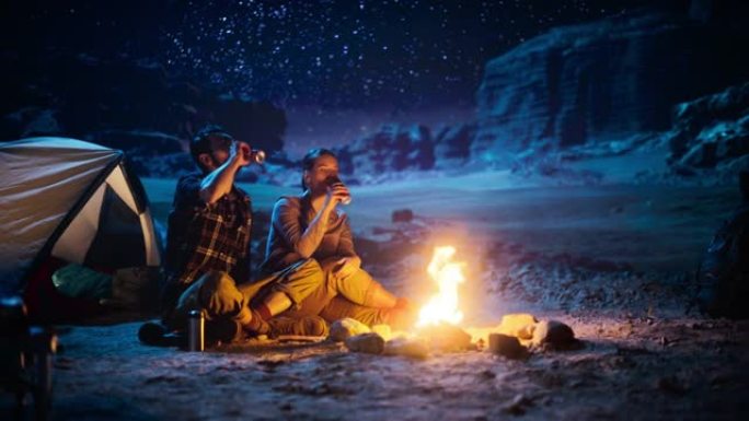 快乐情侣大自然在峡谷露营，坐在篝火旁分享一杯饮料，看夜空。两个旅行的年轻人说话，看看银河系。励志度假