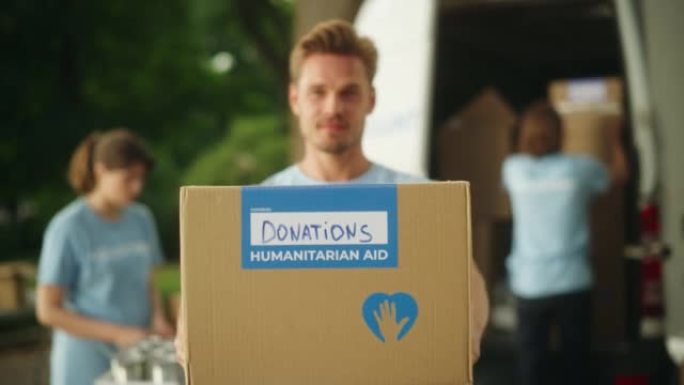 一位英俊的年轻男性志愿者的肖像。穿着蓝色t恤的成功白人男子微笑着，用纸板箱向观众伸出手。人道主义援助