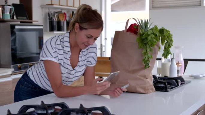 美丽的拉丁美洲女人用智能手机和信用卡在网上购物时，肘部躺在厨房的柜台上