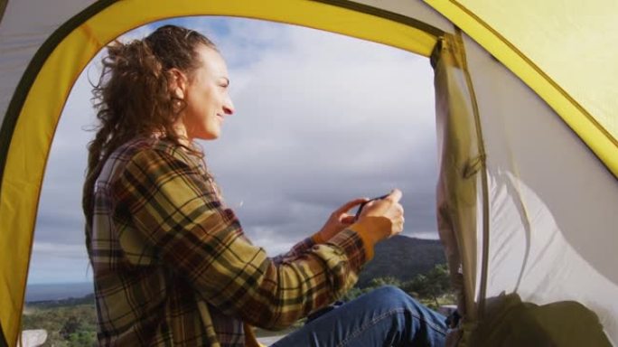 快乐的白人妇女露营，坐在乡村山上的帐篷外面，用智能手机拍照