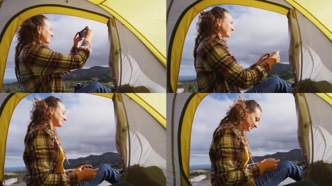 快乐的白人妇女露营，坐在乡村山上的帐篷外面，用智能手机拍照
