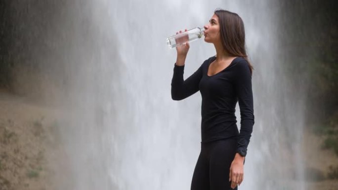 女游客从山上徒步旅行中休息，从瀑布前的水瓶中喝水