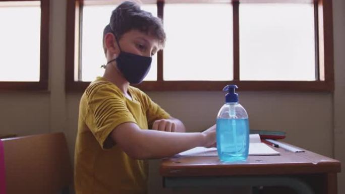 男孩坐在学校的桌子上戴着口罩消毒双手