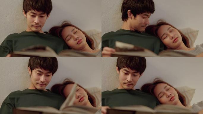 年轻的亚洲男子在晚上女友躺在他的肩膀上看书。