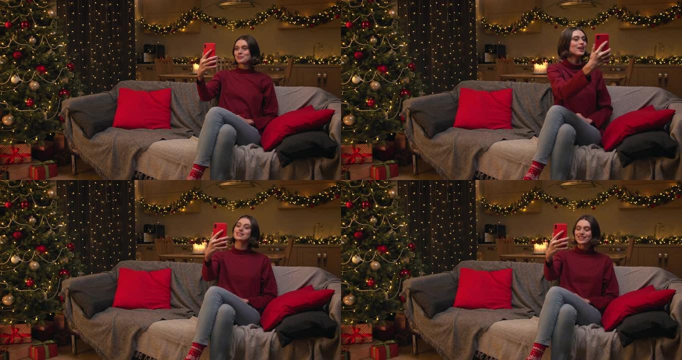 一位穿着红色毛衣的迷人棕色头发的女人正在她的红色智能手机上视频聊天，她坐在装饰有圣诞树的房间里的沙发