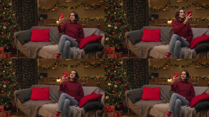 一位穿着红色毛衣的迷人棕色头发的女人正在她的红色智能手机上视频聊天，她坐在装饰有圣诞树的房间里的沙发