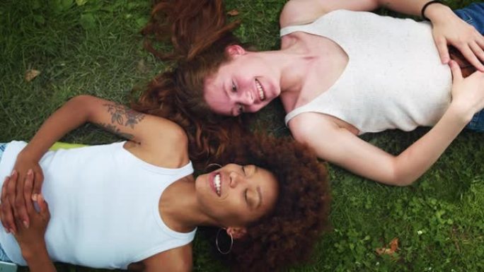 从高处看:两个多民族妇女躺在公园草坪上，有说有笑。不同的女性青少年享受彼此的陪伴，一起在外面度过时光