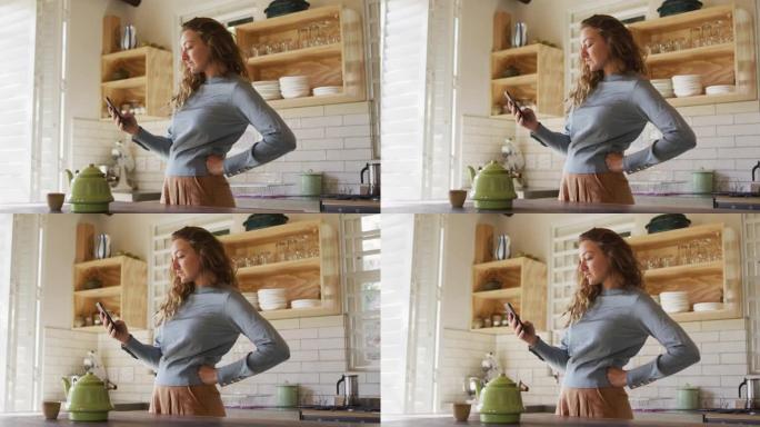 白人妇女用智能手机站在小屋厨房里，用手放在臀部上，茶壶放在柜台上