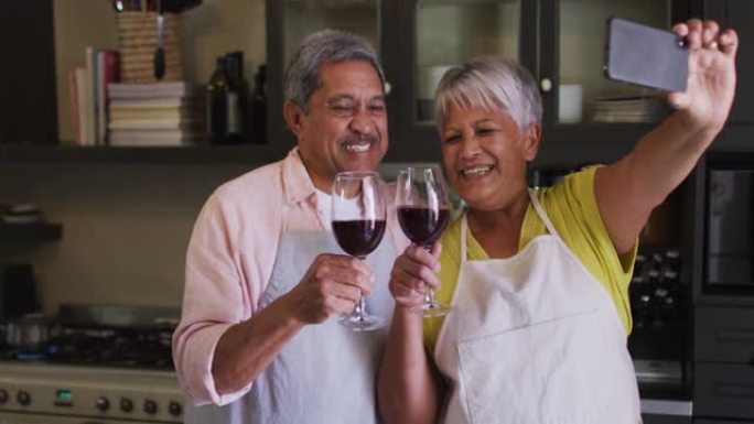 快乐的高级混血夫妇在厨房里用葡萄酒自拍照敬酒