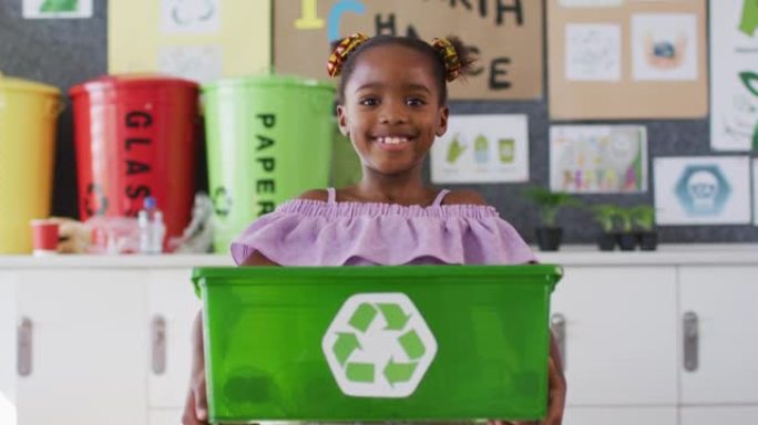 快乐的非洲裔美国女学生站在教室里，微笑着，拿着回收箱