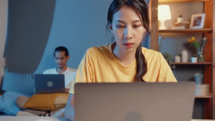 自由职业者亚洲夫妇在休闲妻子专注于财务工作业务晚上在桌上的笔记本电脑上快乐的丈夫视频通话与家人休息在