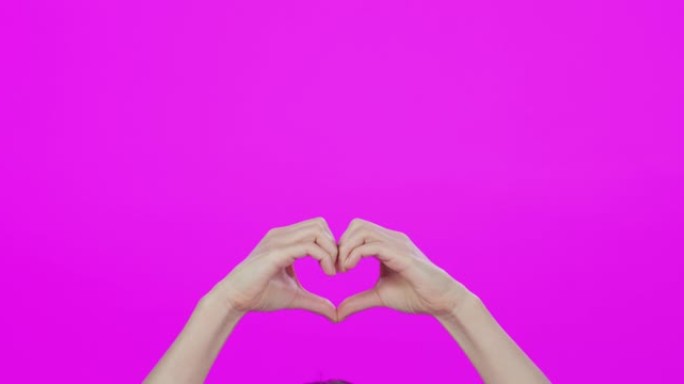 4k视频片段，一个无法识别的女人在粉红色的工作室背景下做出心形手势