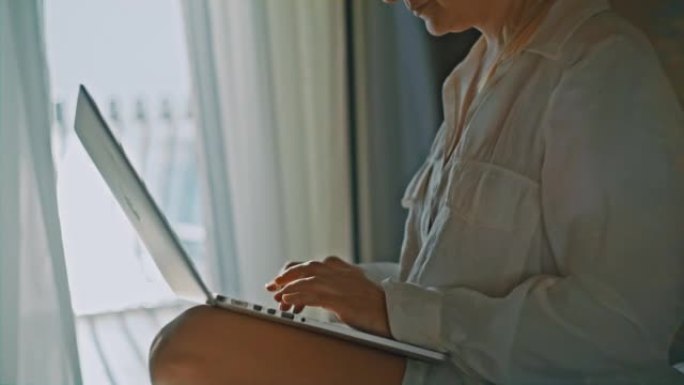 女人坐在酒店房间的床上在笔记本电脑上打字，背景是大海的阳台，透明的窗帘在风中移动