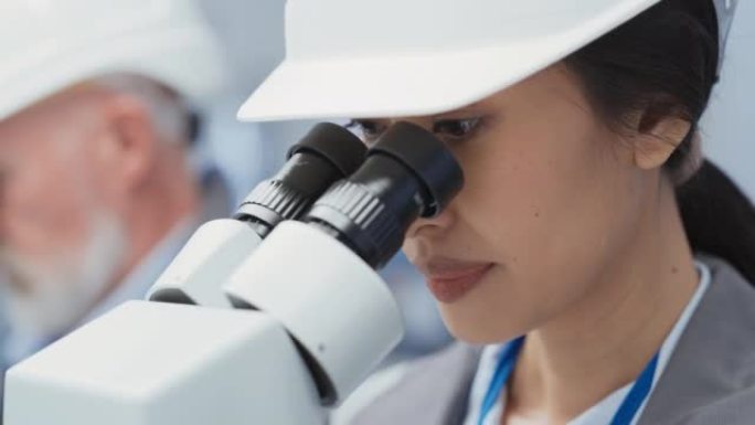 一位美丽的年轻亚洲工业科学家戴着白色安全帽，在工厂的办公桌前工作，使用显微镜检查制造生产零件的质量。