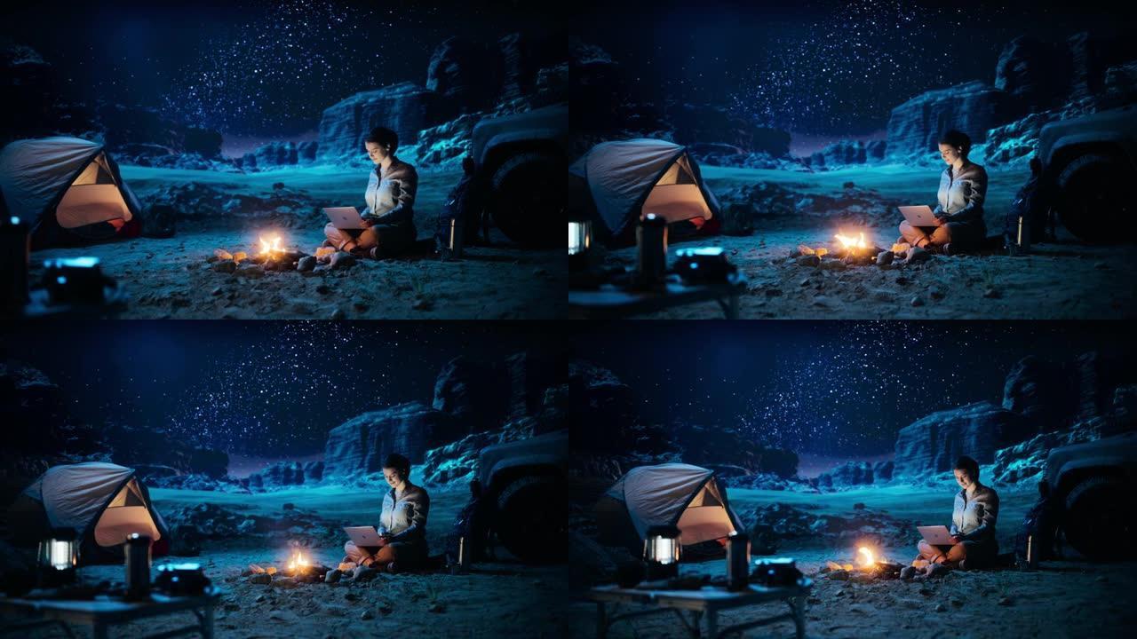 峡谷夜间帐篷露营: 女性旅行者使用坐在篝火旁的笔记本电脑。女性从事数字远程工作，电子购物，电子商务，
