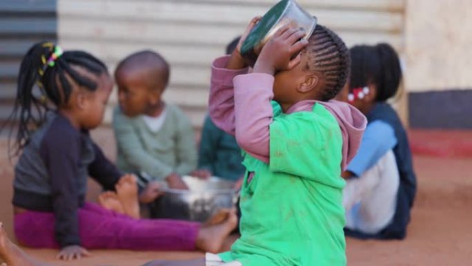 非洲的贫困。年轻的饥饿黑人非洲女孩从锅里吃玉米，三个孩子在背景中