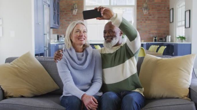 快乐的老年多样化夫妇坐在沙发上，使用智能手机，进行视频通话