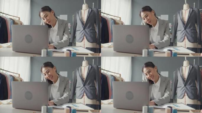 专业开朗的亚洲女商人设计师坐在办公桌上使用笔记本电脑呼叫智能手机，以在工作室工作场所在线市场上确认客